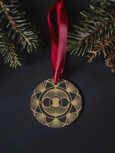 Round metal ornament featuring golden triple moon symbol surrounded by Art Deco sunburst pattern on black background. 3.5-inch diameter piece with symmetrical lunar design and crimson ribbon. Detailed celestial artwork adorns both side
